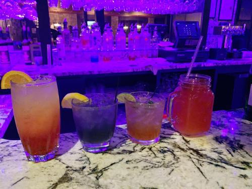 Four colorful cocktails with garnishes sit on a marble bar, illuminated by purple lighting.