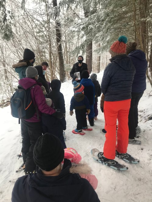 A group of people, including children, gather in a snowy forest for a winter activity, listening to an instructor.