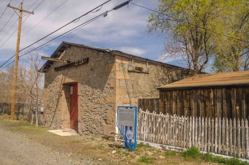 A stone building with a red door, surrounded by a wooden fence and trees, under a blue sky with scattered clouds.