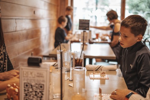 A boy sits at a table in a restaurant, focused on a game, while others are engaged in conversation nearby.