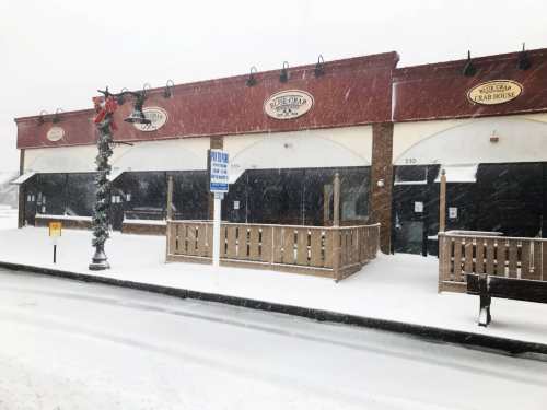 A crab house restaurant covered in snow during a heavy snowstorm, with empty outdoor seating and a street sign visible.