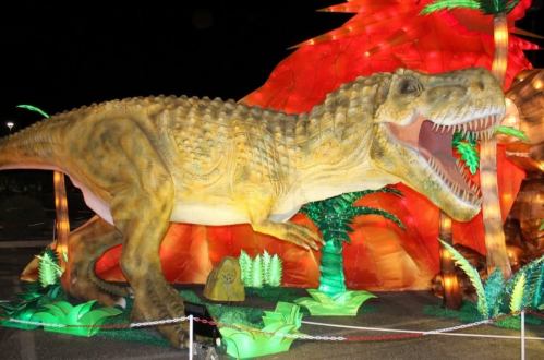 A large, illuminated dinosaur sculpture with a fierce expression, surrounded by colorful foliage and a vibrant backdrop.