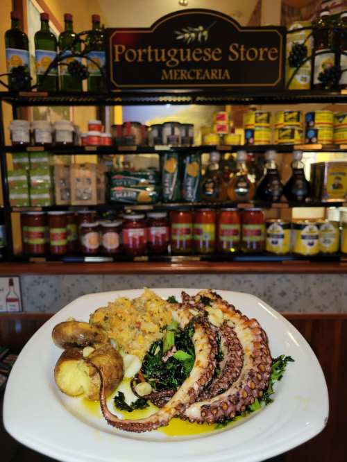 A plate of octopus with potatoes and greens, set against a backdrop of shelves filled with various canned goods.