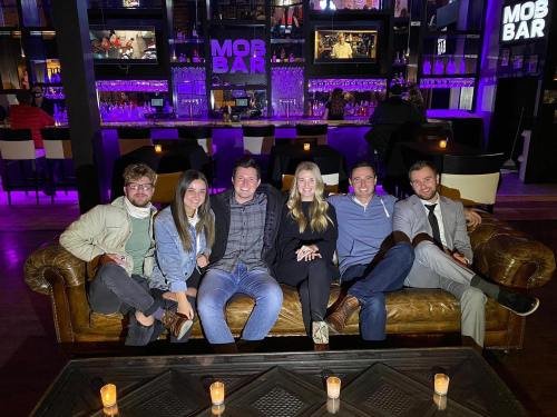 A group of six friends sitting on a leather couch in a dimly lit bar with purple lighting and candles on the table.