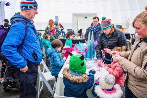 A group of children and adults engage in a craft activity inside a large, bright tent during a festive event.