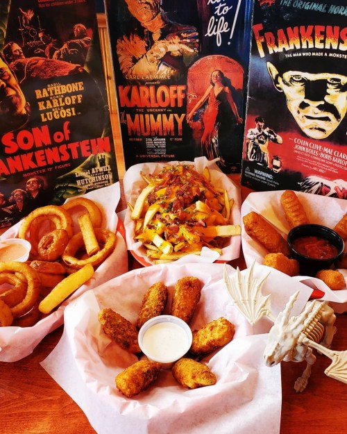 A spread of fried snacks including onion rings, fries topped with chili, and mozzarella sticks, with classic horror movie posters in the background.