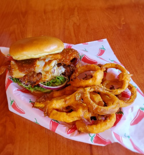 A delicious burger with melted cheese and toppings, served with crispy onion rings on a red chili-patterned paper.