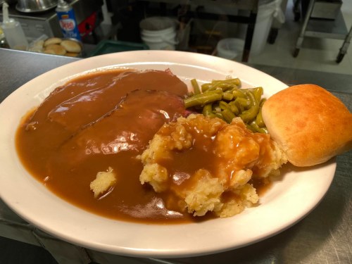 A plate of meat with gravy, mashed potatoes, green beans, and a roll, served in a diner setting.