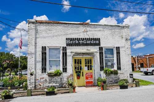 A quaint white brick building with a yellow door, labeled "Herb Garden," surrounded by plants and an "Open" sign.