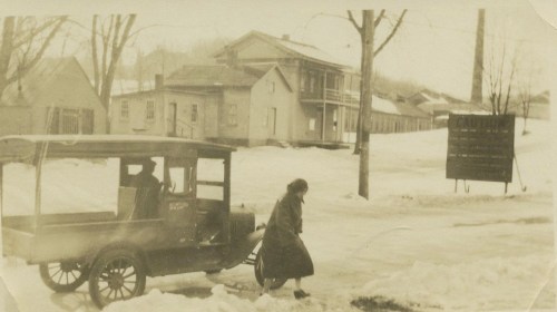 A vintage photo of a woman walking near a snow-covered street with an old truck and buildings in the background.