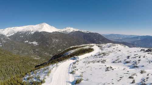 A snowy mountain landscape with a winding road and a vehicle, surrounded by tall trees and distant peaks under a clear sky.