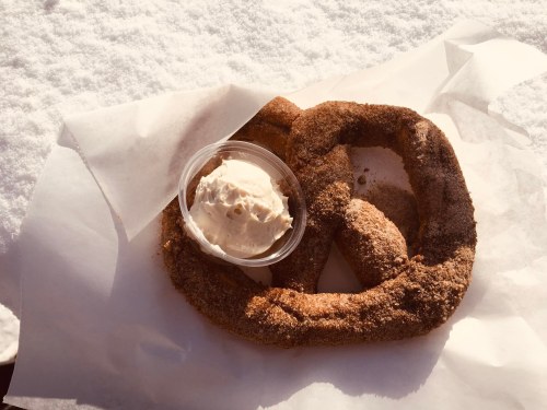 A large cinnamon sugar pretzel on parchment paper, accompanied by a small cup of cream cheese frosting.