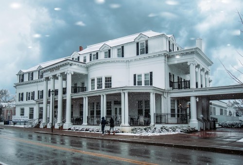 A large white building with columns, surrounded by snow, under a cloudy sky. Two people walk on the street in front.