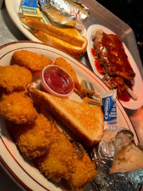 A plate of crispy chicken tenders, toast, and a baked potato with sour cream, alongside a BBQ rib platter and bread.