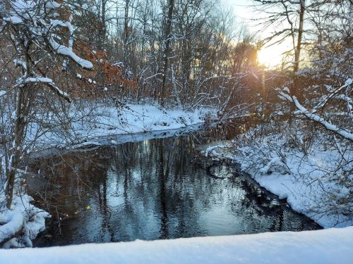 A serene winter scene with a snow-covered river surrounded by trees, reflecting the warm glow of the setting sun.