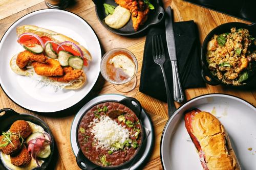 A spread of various dishes including sandwiches, fried chicken, beans, and a salad on a wooden table.