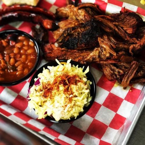 A plate of barbecue featuring smoked meat, baked beans, and coleslaw on a red and white checkered tray.