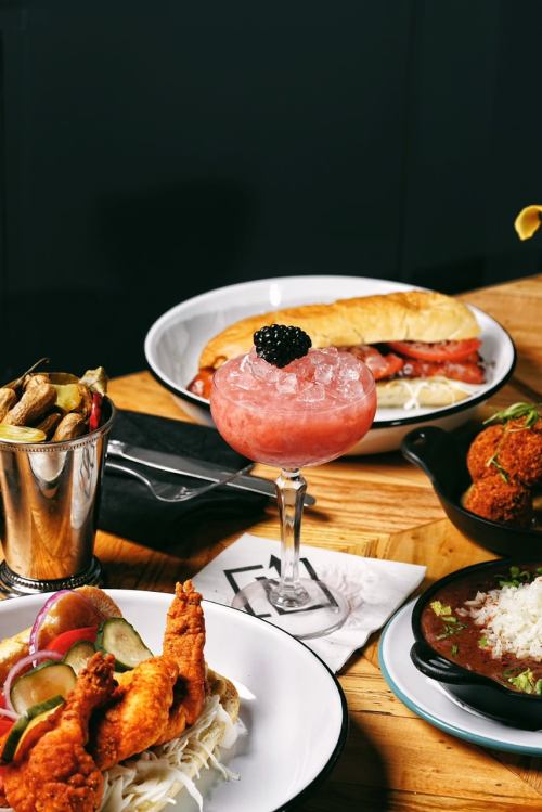 A table set with a cocktail, fried chicken sandwich, and various side dishes, all on wooden surfaces.