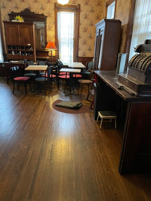 A cozy room with wooden floors, vintage furniture, and a mix of tables and chairs, illuminated by natural light.