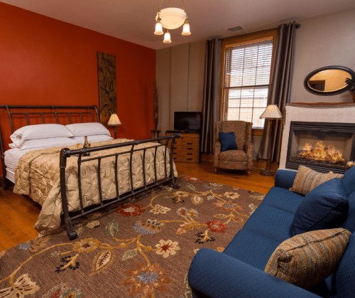 Cozy bedroom with a metal bed, fireplace, armchair, and a patterned rug on wooden floors. Warm orange wall.