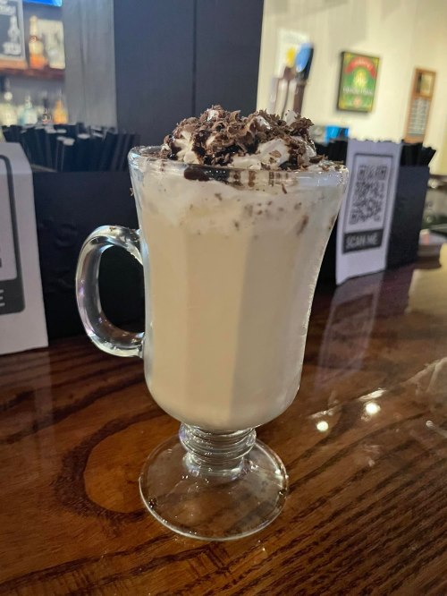 A creamy dessert drink topped with whipped cream and chocolate shavings, served in a glass on a wooden bar.