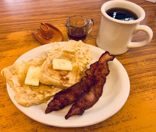 A plate with French toast topped with butter, crispy bacon, a small syrup cup, and a coffee mug on a wooden table.