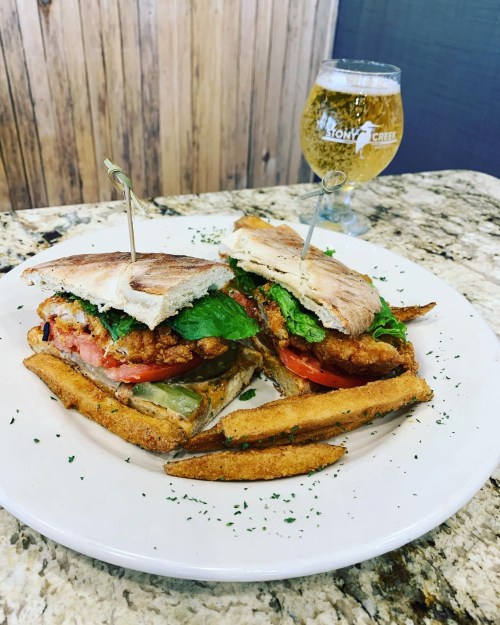 A delicious sandwich with fried chicken, lettuce, and tomato, served with fries and a glass of beer on the side.