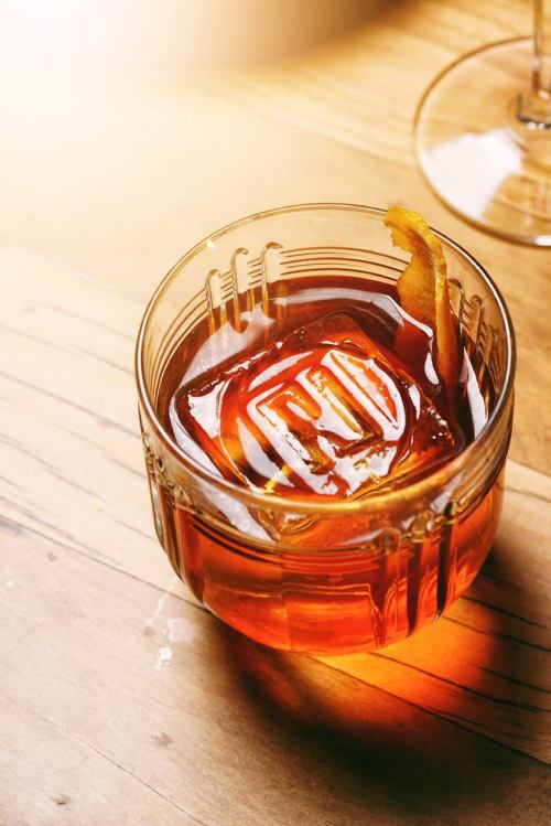 A glass of amber-colored cocktail with a large ice cube and a twist of citrus peel on a wooden surface.