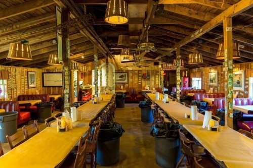 Interior of a rustic dining hall with wooden beams, long tables, and trash bins, featuring cozy seating and warm lighting.