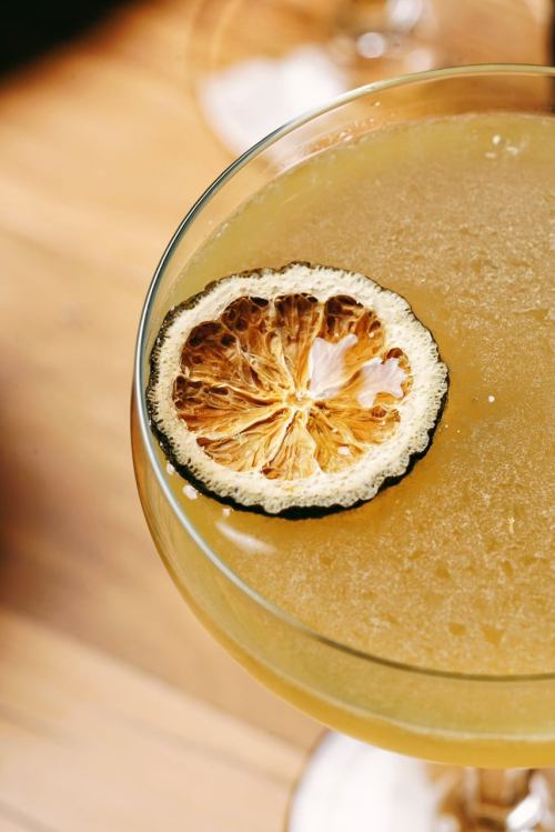A close-up of a cocktail in a glass, topped with a dried lemon slice on a wooden surface.