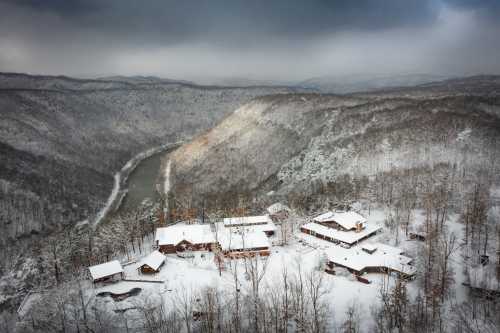 Aerial view of a snowy landscape with a river and rustic buildings nestled among trees on a mountain.