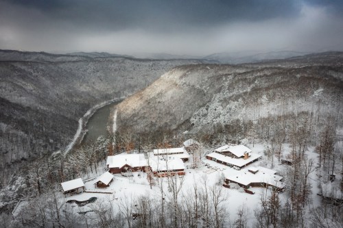 Aerial view of a snowy landscape with a river and rustic buildings nestled among trees on a mountain.