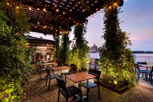 Outdoor dining area with wooden tables, surrounded by greenery and string lights, overlooking a river at sunset.