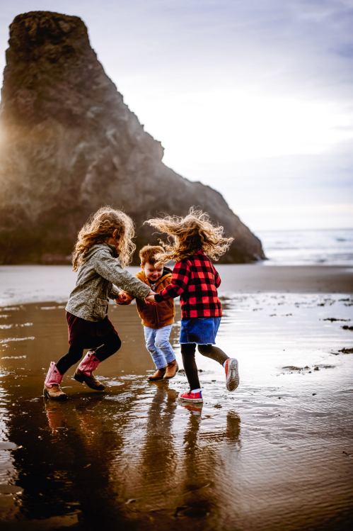 Three children with curly hair play joyfully on a beach, splashing in the water with a rocky cliff in the background.