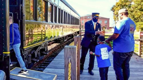 A conductor hands tickets to a family near a vintage train at a station on a sunny day.