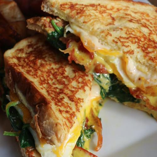 A close-up of a grilled cheese sandwich with spinach, bacon, and melted cheese, served with crispy potato wedges.