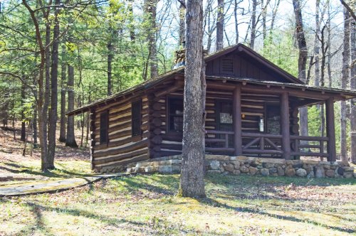 A rustic log cabin surrounded by trees in a wooded area, featuring a stone foundation and a porch.