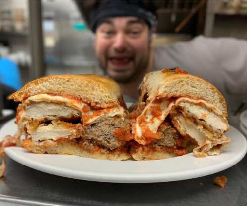 A close-up of a hearty sandwich filled with meat and sauce, with a smiling chef in the background.