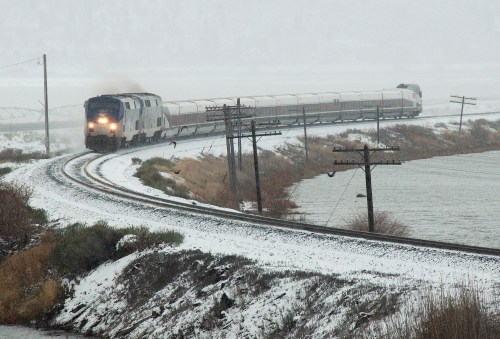 A train curves along snowy tracks beside a body of water, with snow-covered hills in the background.