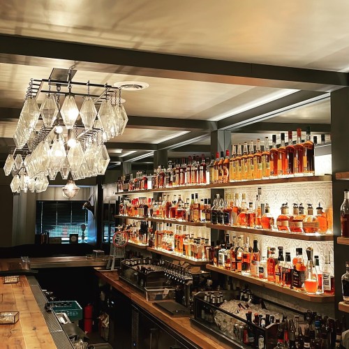 A stylish bar with shelves filled with various bottles of liquor, illuminated by a modern chandelier.