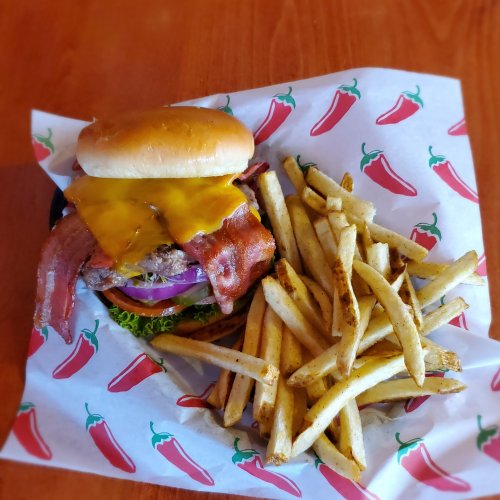 A cheeseburger with bacon, lettuce, and onion, served with a side of golden fries on a chili-patterned paper.