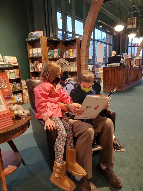 Two children read a book together while sitting on a man's lap in a cozy bookstore.