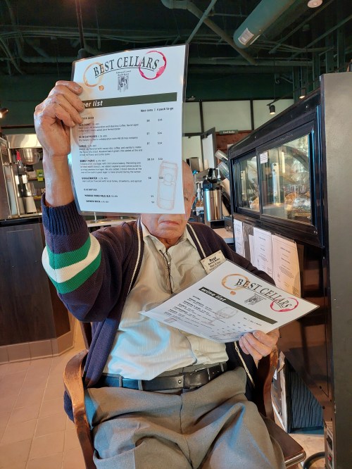 An elderly man sits in a café, holding two menus while looking at one, with a cozy interior in the background.