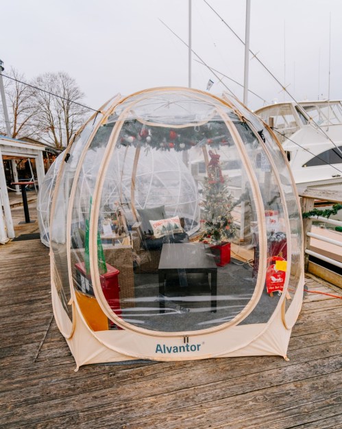 A transparent outdoor igloo tent on a dock, decorated for the holidays with a small Christmas tree and festive decor inside.
