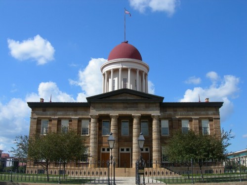 Historic building with a red dome, columns, and flag, surrounded by trees and blue sky with clouds.