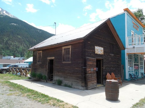 A rustic wooden building with a metal roof, surrounded by greenery and mountains in the background.