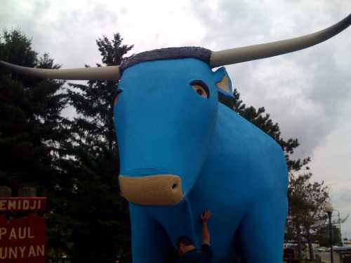 A large blue statue of a bull with prominent horns, set against a backdrop of trees and a cloudy sky.