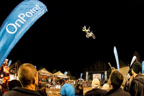 A motocross rider performs a jump at night, with spectators watching and banners in the foreground.