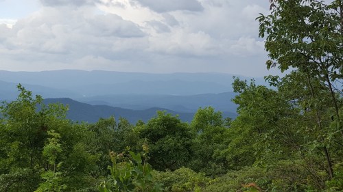 A scenic view of layered mountains under a cloudy sky, framed by lush green trees in the foreground.