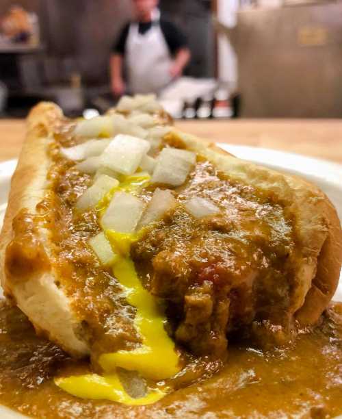 A close-up of a hot dog topped with chili, mustard, and diced onions, with a blurred kitchen background.